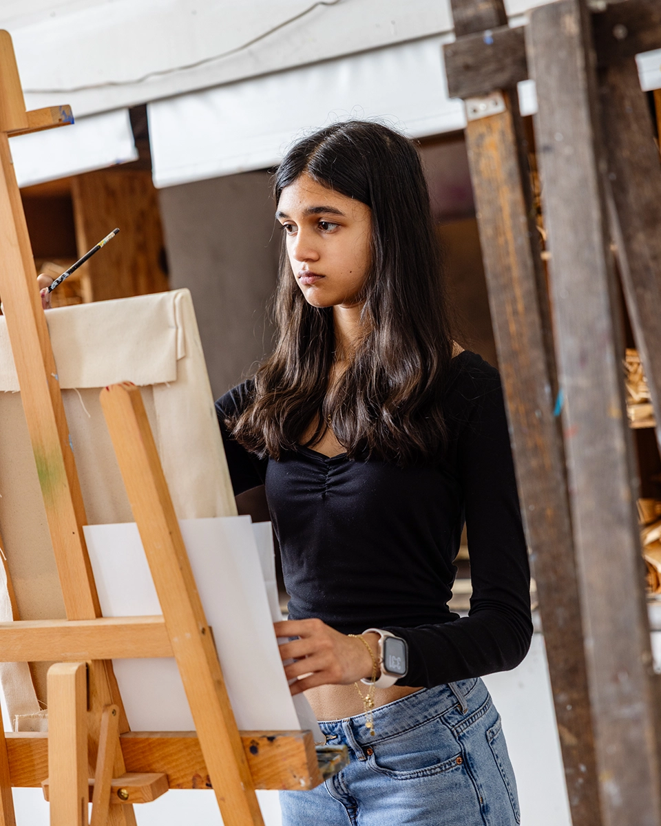 A young woman with long dark hair, wearing a black top and jeans, paints on a canvas at an easel in an art studio, holding a brush and focused on her work.