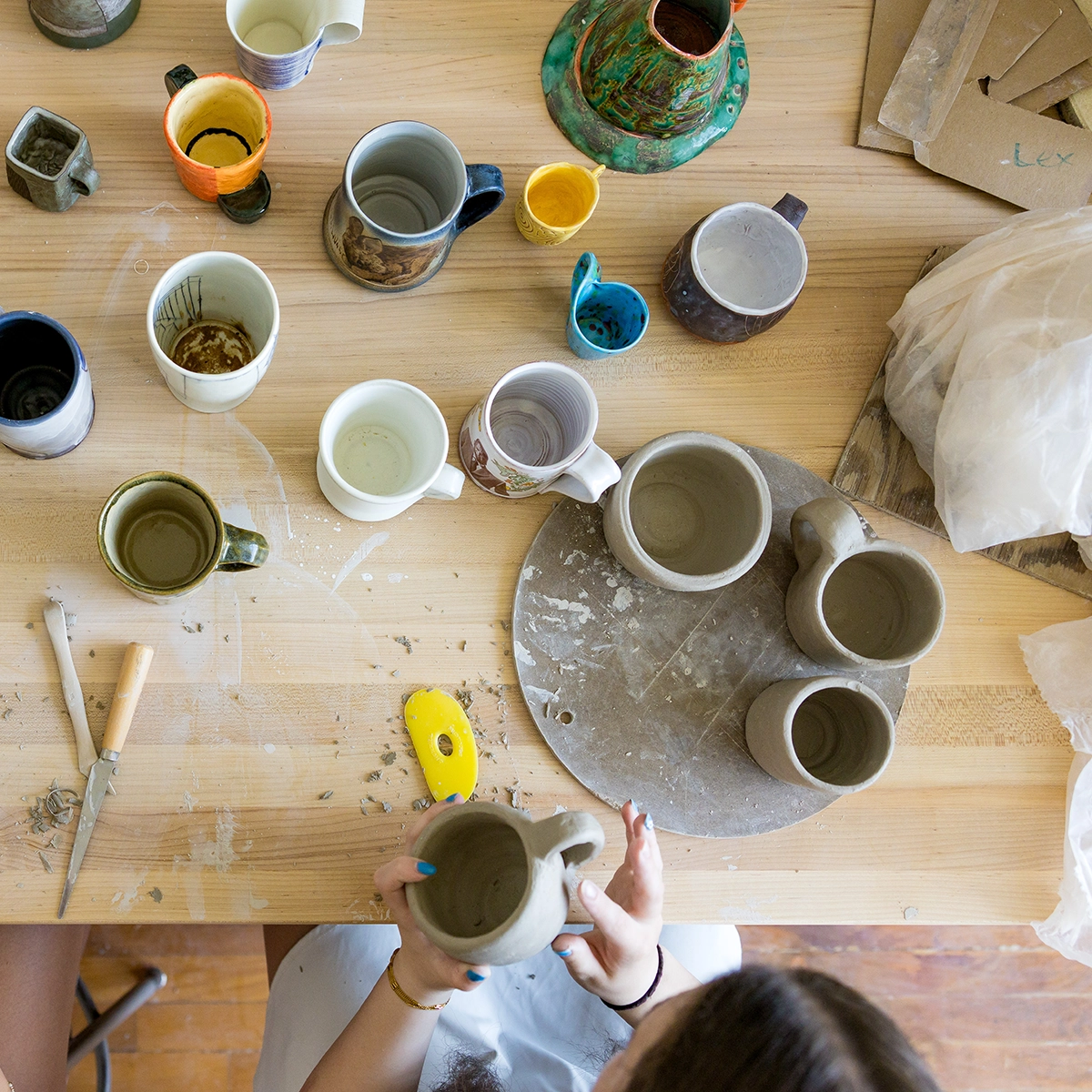 Two people work at a wooden table filled with ceramic mugs, pottery tools, and clay, viewed from above. One person holds an unfinished mug while the other’s hands are just visible at the table’s edge.