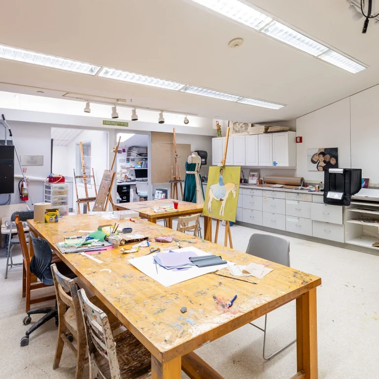 A bright art studio with large tables, easels, scattered art supplies, cabinets, and a computer. The room is well-lit with fluorescent lights and has various chairs and storage areas.