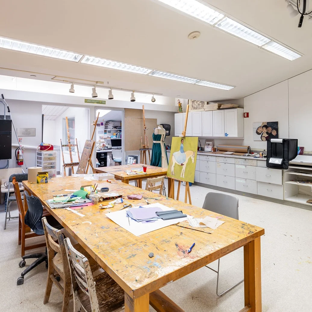 A bright art studio with large tables, easels, scattered art supplies, cabinets, and a computer. The room is well-lit with fluorescent lights and has various chairs and storage areas.