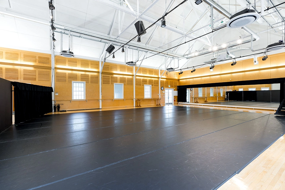 Spacious dance studio with a black floor, large mirrors along one wall, high white ceiling with exposed beams, and yellow walls with several windows. Ceiling lights and speakers are also visible.
