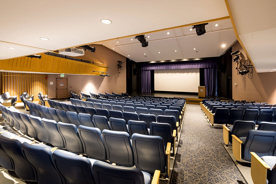 Empty auditorium with rows of blue seats facing a stage with a podium and large projection screen, under ceiling lights, and carpeted floor.