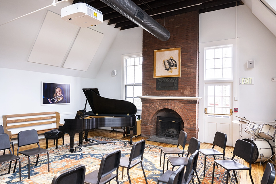 A music room with a grand piano, chairs arranged in a semi-circle, a drum set, a fireplace, portraits on the walls, and large windows letting in natural light.