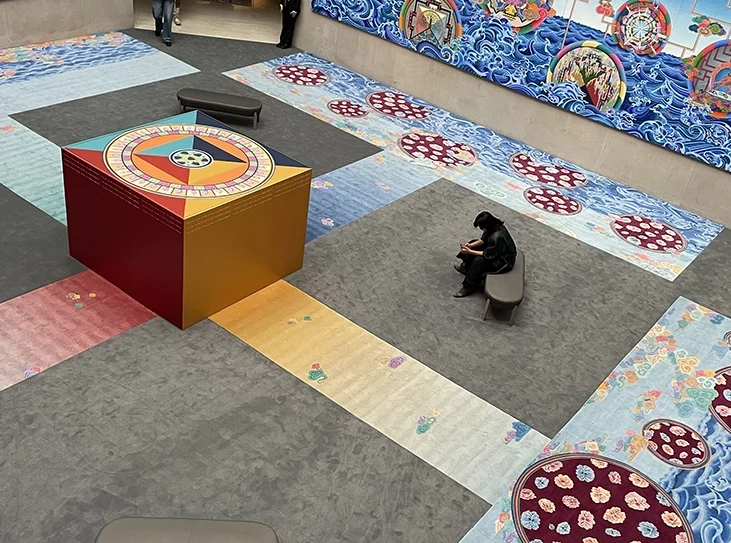 Colorful art installation in a large indoor museum space with patterned carpets, a central square structure, wall tapestries, and people seated or walking around. The scene blends vibrant colors and intricate designs.