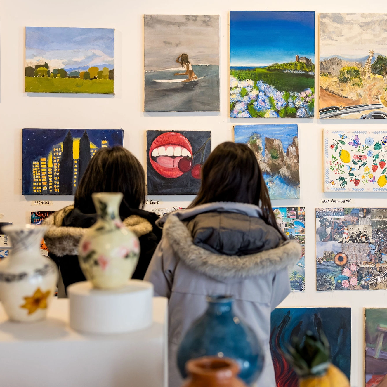 Two people in winter coats view colorful paintings displayed on a gallery wall. Painted vases sit on a white pedestal in the foreground, partly obscuring the view of the people and artwork.