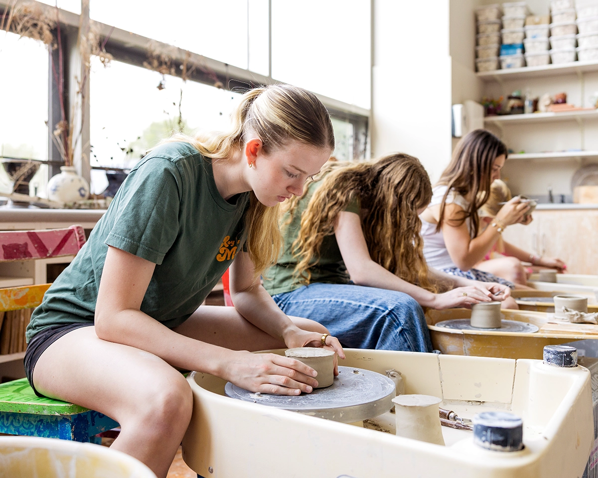 Three young women sit at pottery wheels in a bright studio, shaping clay with their hands. Shelves with supplies and large windows are visible in the background.