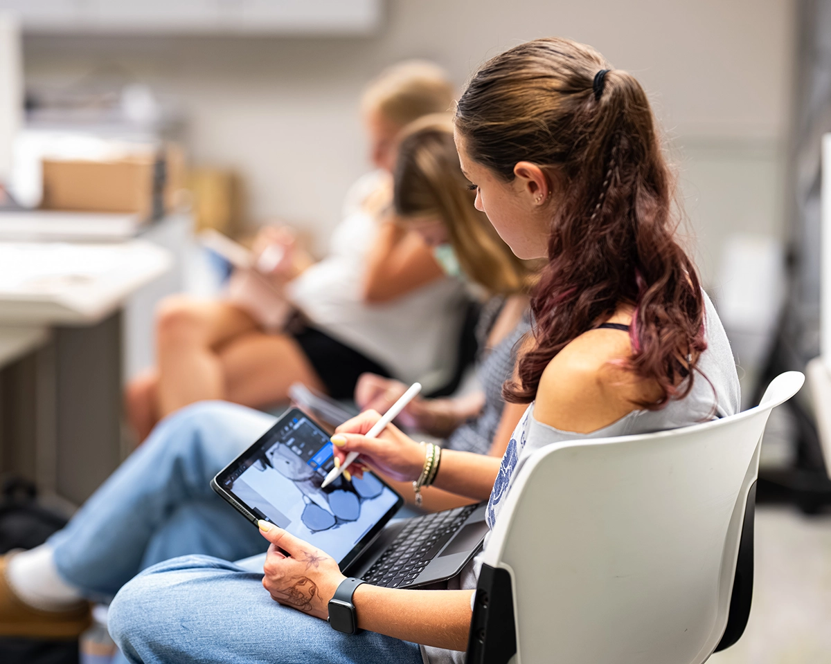 A woman sits in a chair using a tablet and stylus to draw, with others seated and working in the background. The setting appears to be a classroom or workshop.
