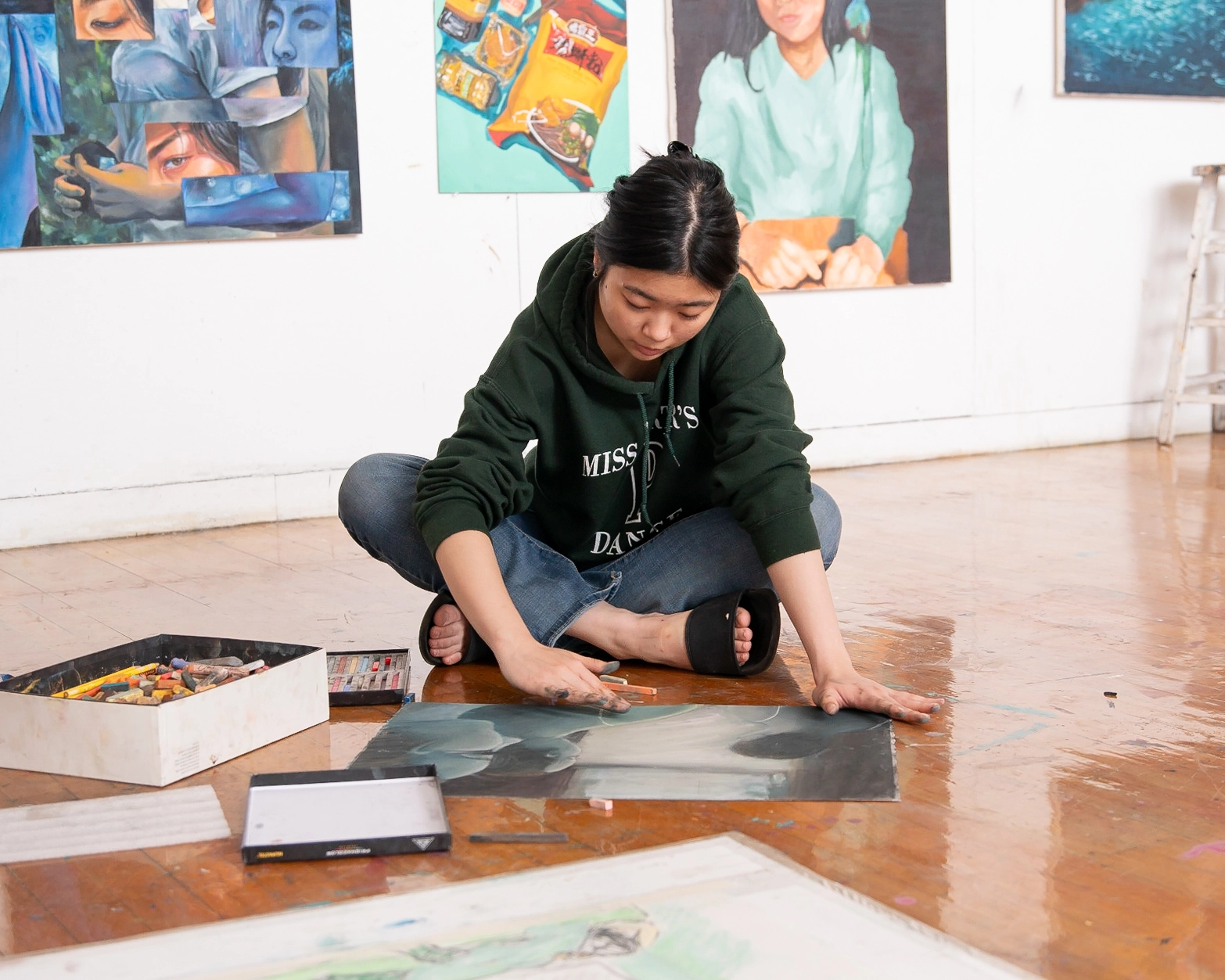 A woman sits on a wooden floor, drawing with pastels on large paper. Art supplies and finished artworks surround her in a bright studio with paintings hanging on the walls.