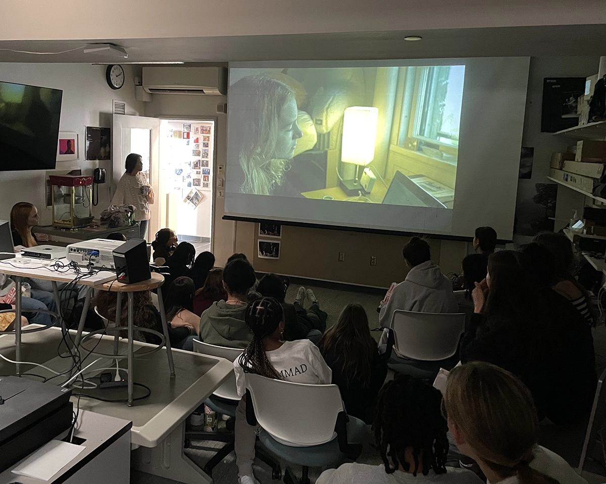 A group of students sits in a dimly lit classroom watching a film projected on a screen, featuring a close-up of a woman by a lamp. The room has chairs, desks, and various equipment visible.