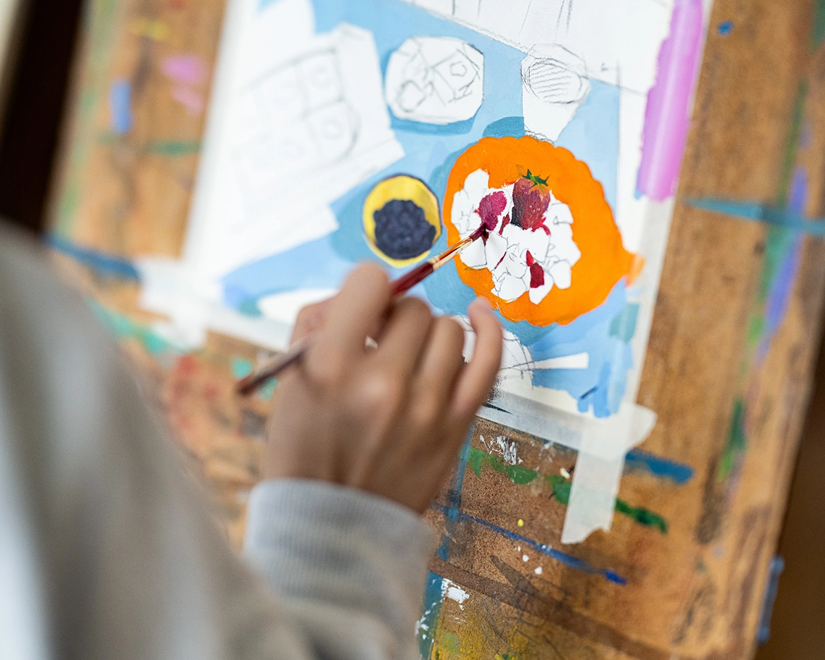 A person paints a colorful still life on canvas, focusing on a plate of fruit using a fine brush. The painting is vibrant, with visible blue, orange, and white areas on a paint-splattered wooden surface.