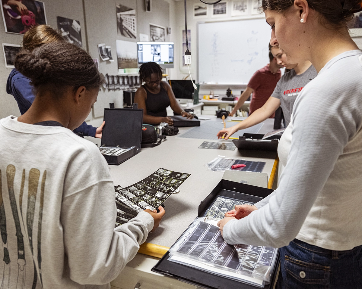 Several people work at a table in a classroom, examining and organizing black-and-white photographic film negatives in plastic sleeves and boxes. Photographs and art supplies are visible in the background.