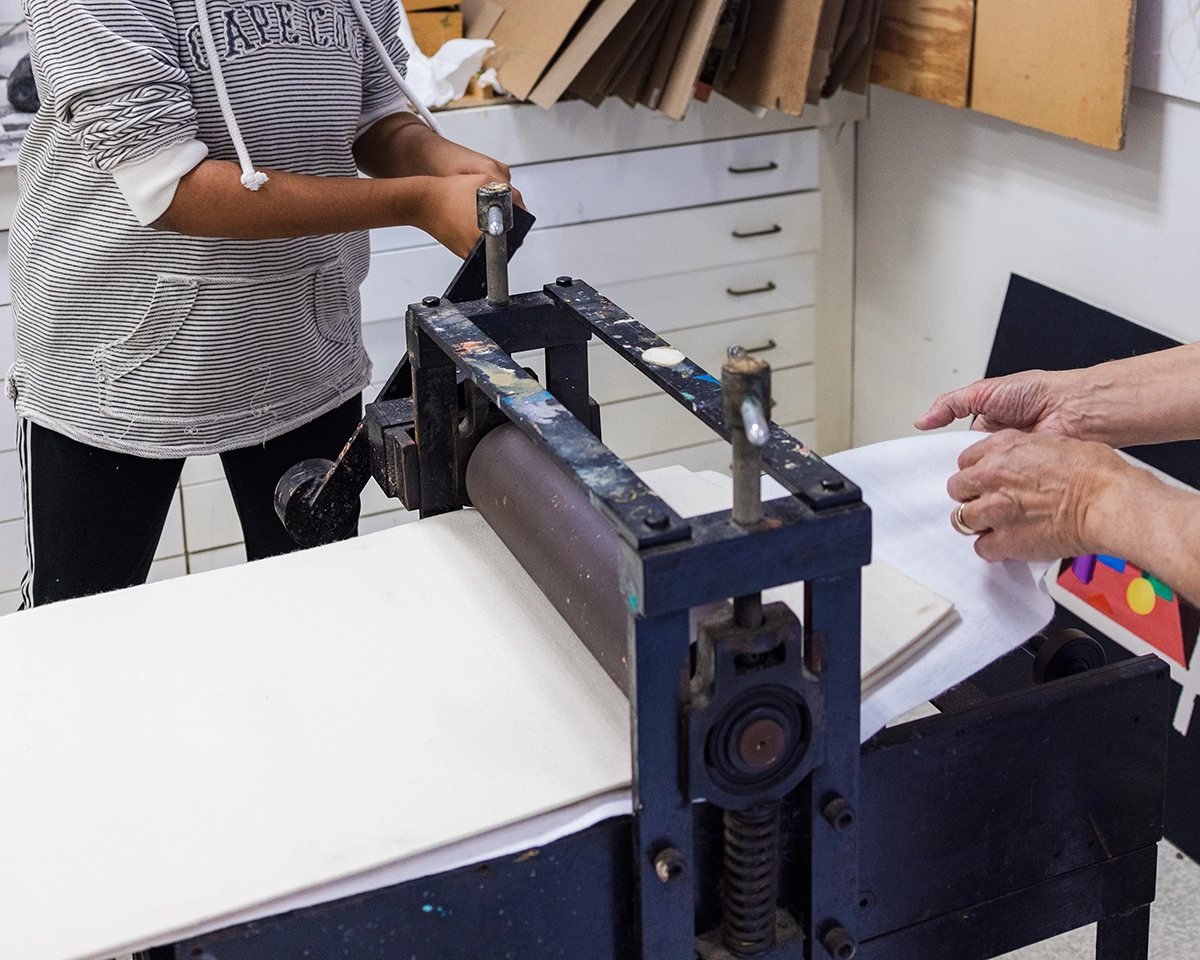 Two people use a printmaking press in an art studio. One turns a handle while the other adjusts paper on the press. Art supplies and paper are visible in the background.