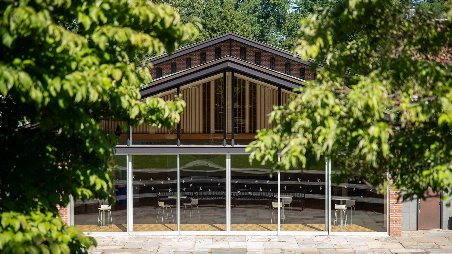 A modern glass building with a peaked roof is partially obscured by green trees. Inside, barstools line a curved counter, and sunlight brightens the open, airy space.