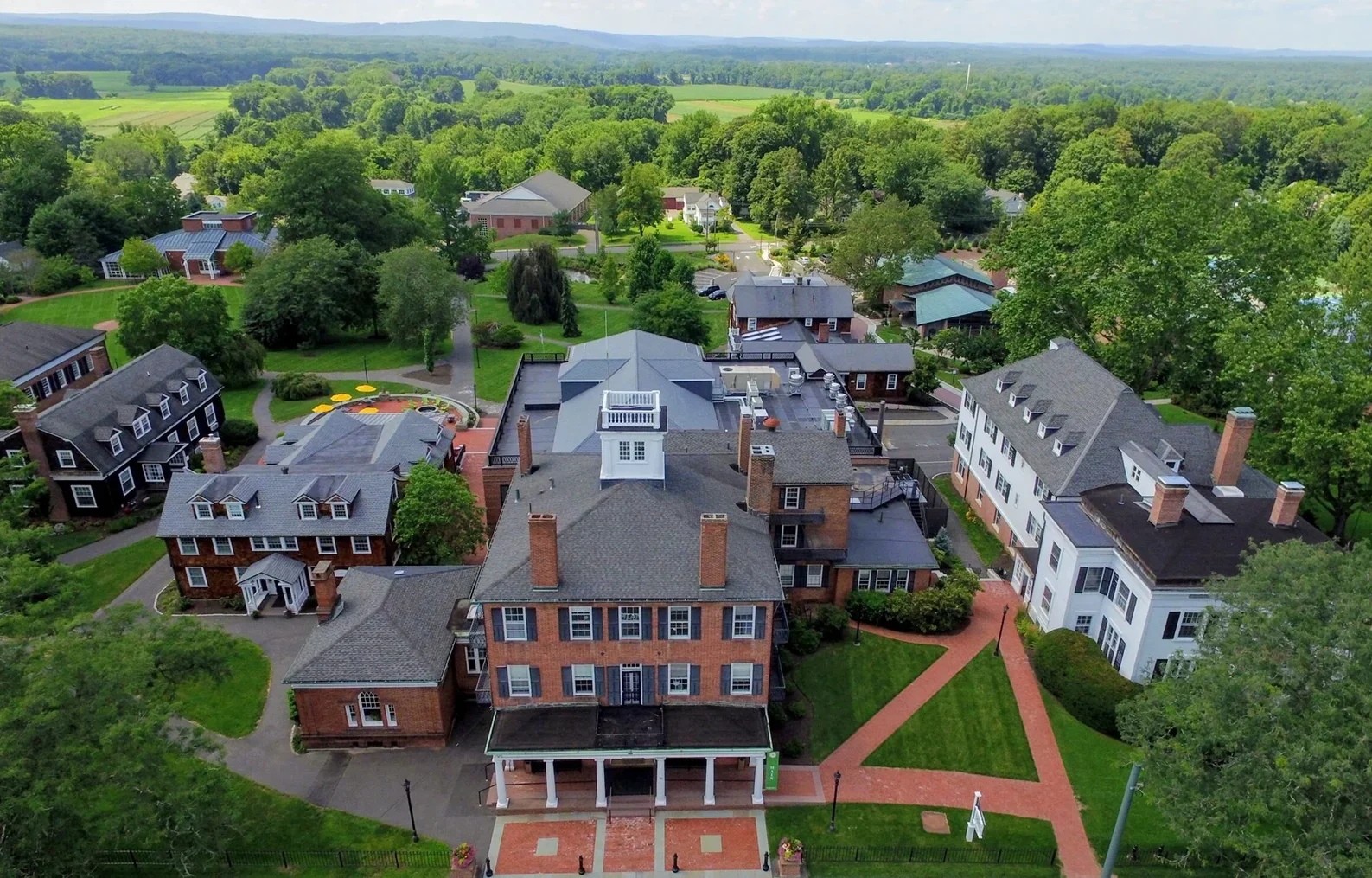 Aerial view of Miss Porter's School campus with historic brick and white buildings, green lawns, pathways, and surrounding trees, set against a backdrop of fields and distant hills under a partly cloudy sky.
