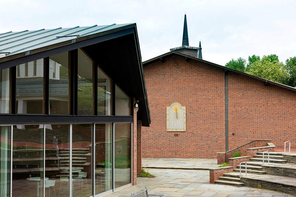 Modern brick and glass building with large windows and a slanted roof, next to a red brick wall featuring a sunburst design. A church steeple and greenery are visible in the background.