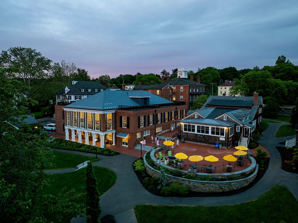 Aerial view of a large brick building with many windows, surrounded by greenery. An outdoor patio area with yellow umbrellas and tables is in the foreground, and paths wind through the landscaped grounds at dusk.