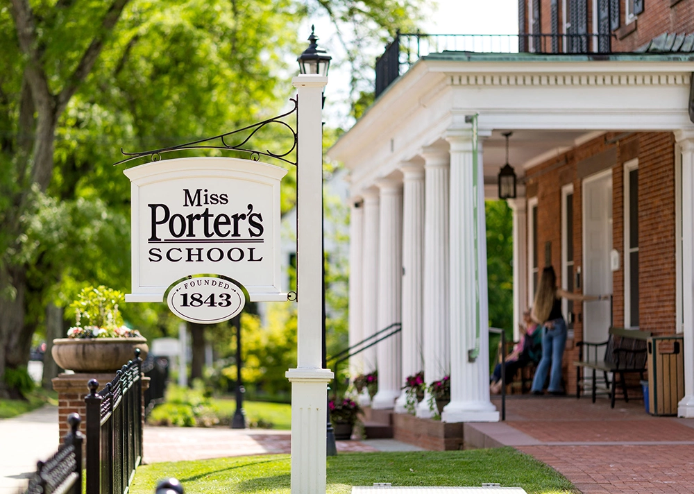 A white sign reading Miss Porter's School Founded 1843 hangs near a brick building with white columns. People are visible on the porch, and trees line the background on a bright day.