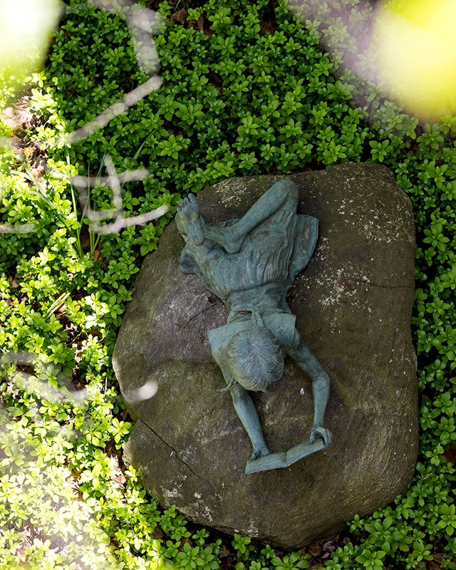 A bronze statue of a child reading a book lies on a large rock, surrounded by dense green ground cover plants, with sunlight filtering through leaves above.