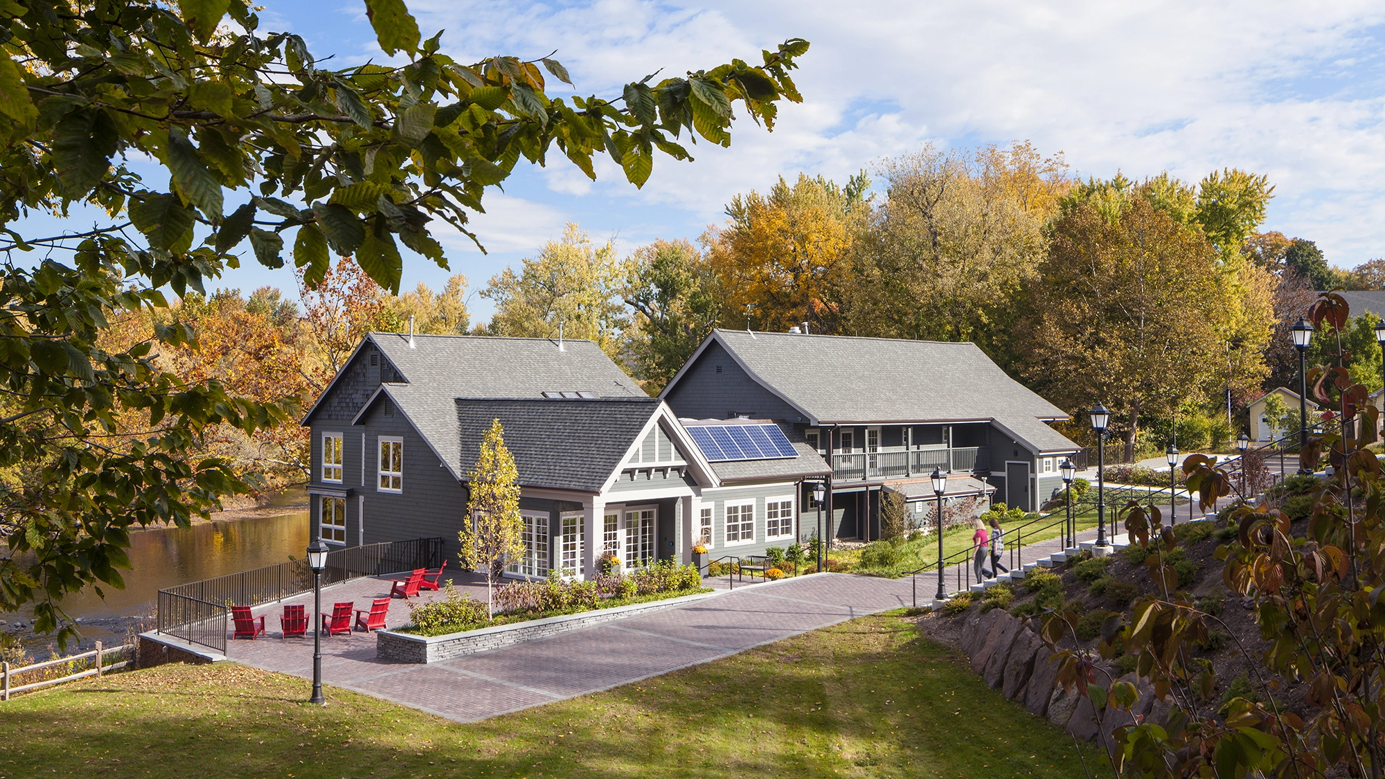 Two gray houses with solar panels sit by a river, surrounded by autumn trees. Red chairs are arranged on a patio, and people walk along a path. Blue sky with scattered clouds is visible above.