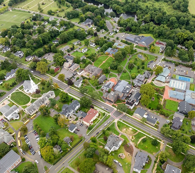 Aerial view of a small town with tree-lined streets, houses, a church, parking lots, sports fields, and surrounding green fields and wooded areas. Roads intersect near the center of the image.