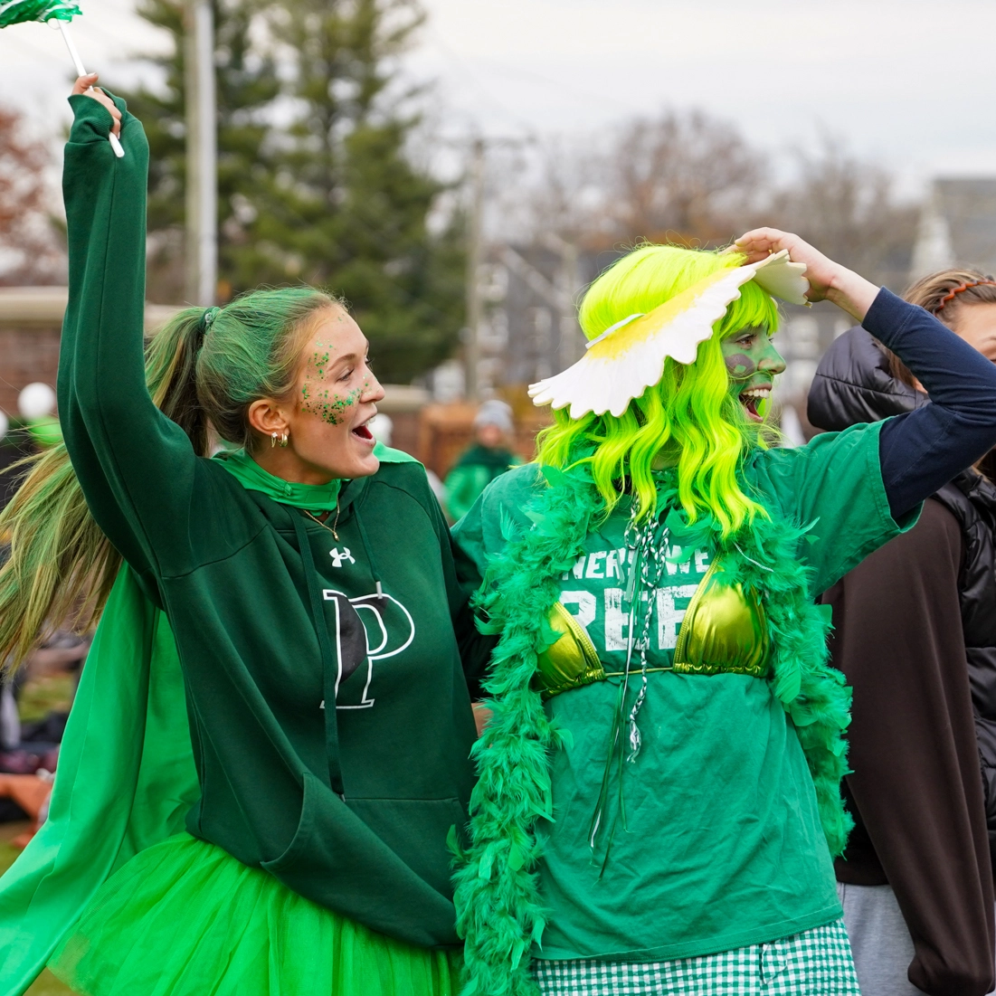 Two people dressed in green with festive accessories and face paint celebrate outdoors, smiling and waving. One wears a tutu and hoodie, the other a yellow wig, feather boa, flower hat, and green checkered skirt.
