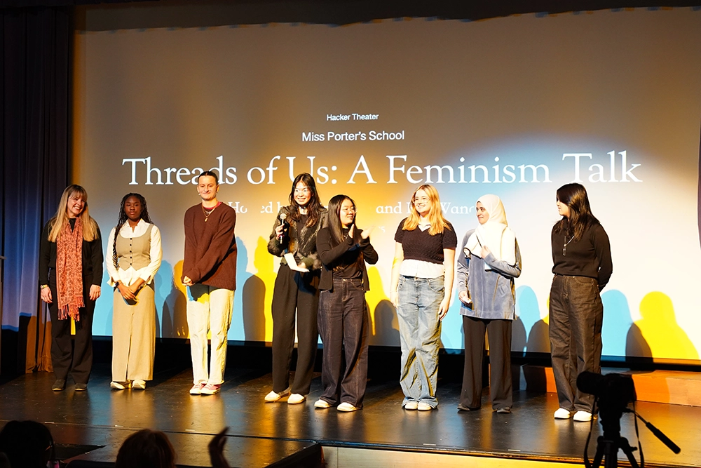 Eight people stand on stage under a screen displaying “Threads of Us: A Feminism Talk.” Some clap and smile as they face the audience, with warm lighting highlighting the group.