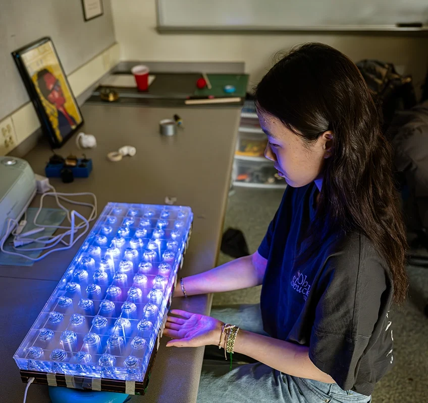 A person examines a large, illuminated keyboard-like device with glowing blue and purple lights in a workspace, surrounded by tools and electronics.