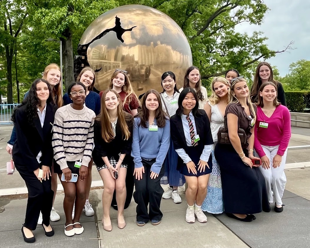 A group of smiling young women pose outdoors in front of a large golden sphere sculpture, with trees and a building in the background. They wear a mix of skirts, dresses, and blouses, and appear happy and cheerful.