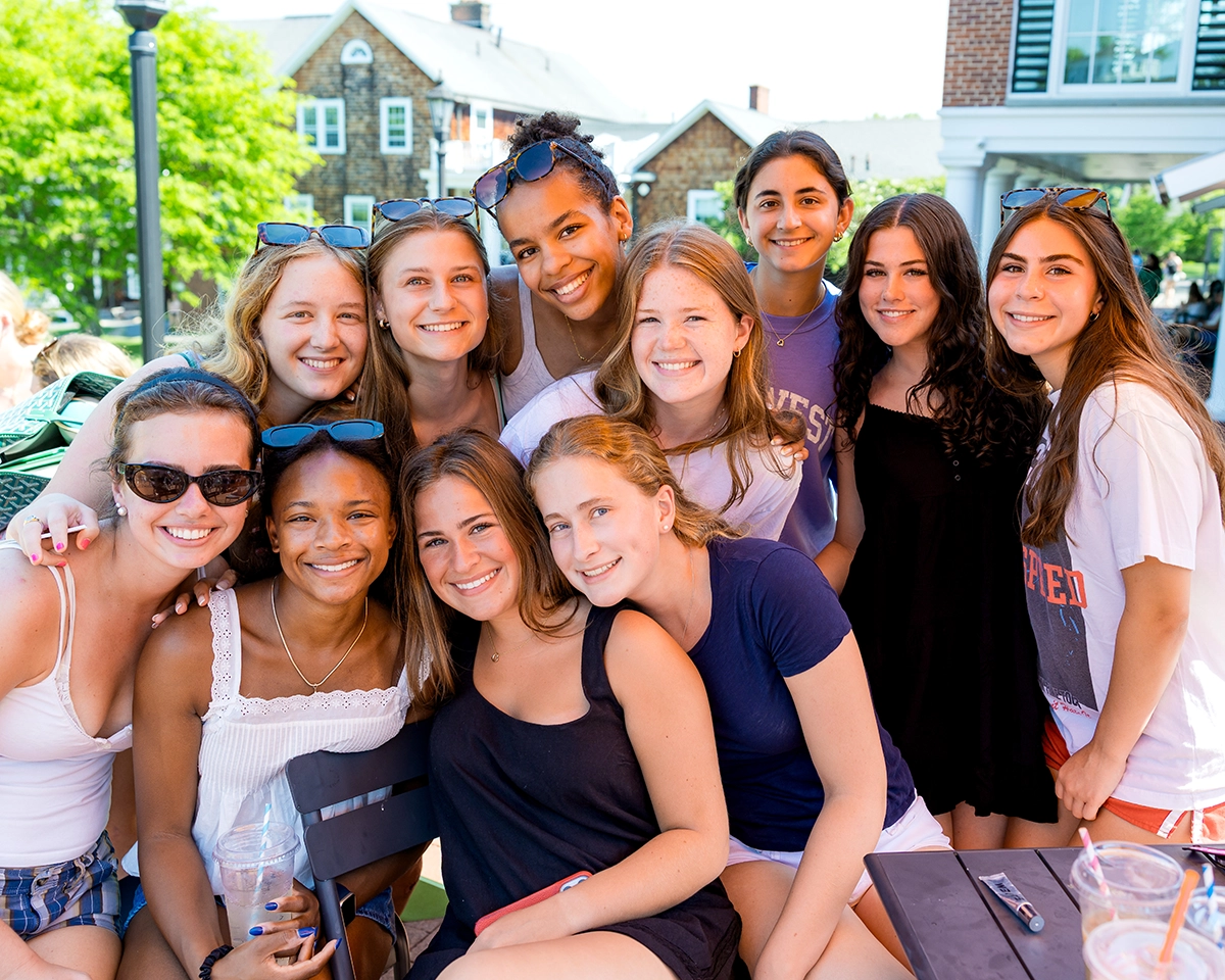 A group of twelve smiling young women pose closely together outdoors on a sunny day, with buildings and greenery visible in the background.