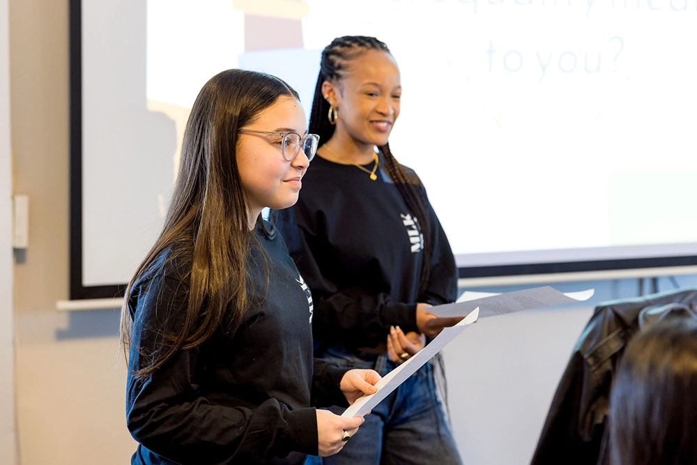 Two young women stand in front of a screen, holding papers and presenting to an audience. Both wear black shirts, and one has glasses while the other has braids, smiling as they speak.