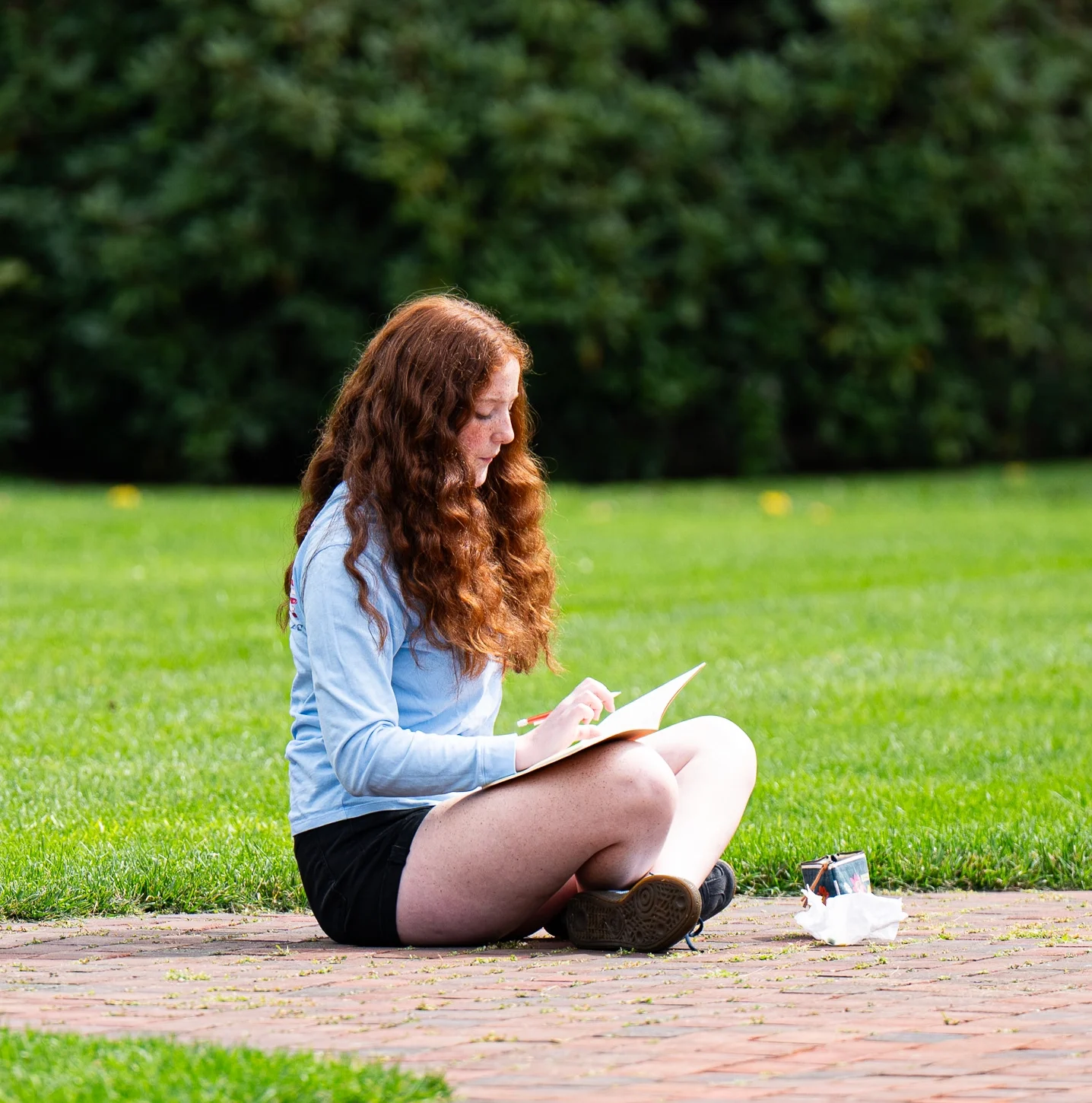 A person with long, curly red hair sits on a brick path in a grassy area, drawing or writing in a notebook on their lap. They wear a light blue shirt and black shorts.