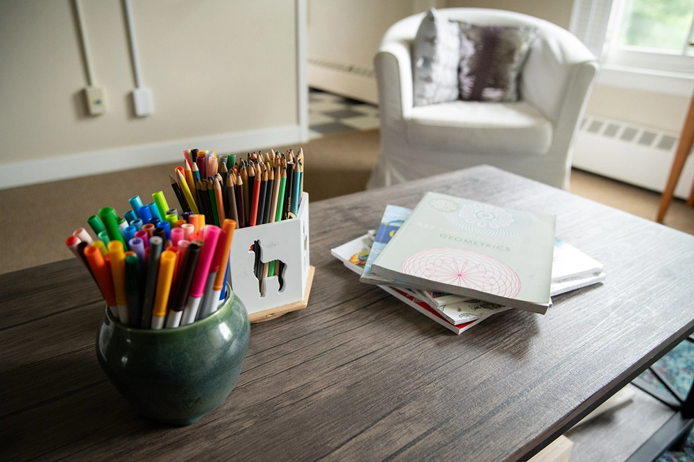 A table with colored pens in a green pot, colored pencils in a holder, and a stack of books. In the background, there is a white armchair by a window.
