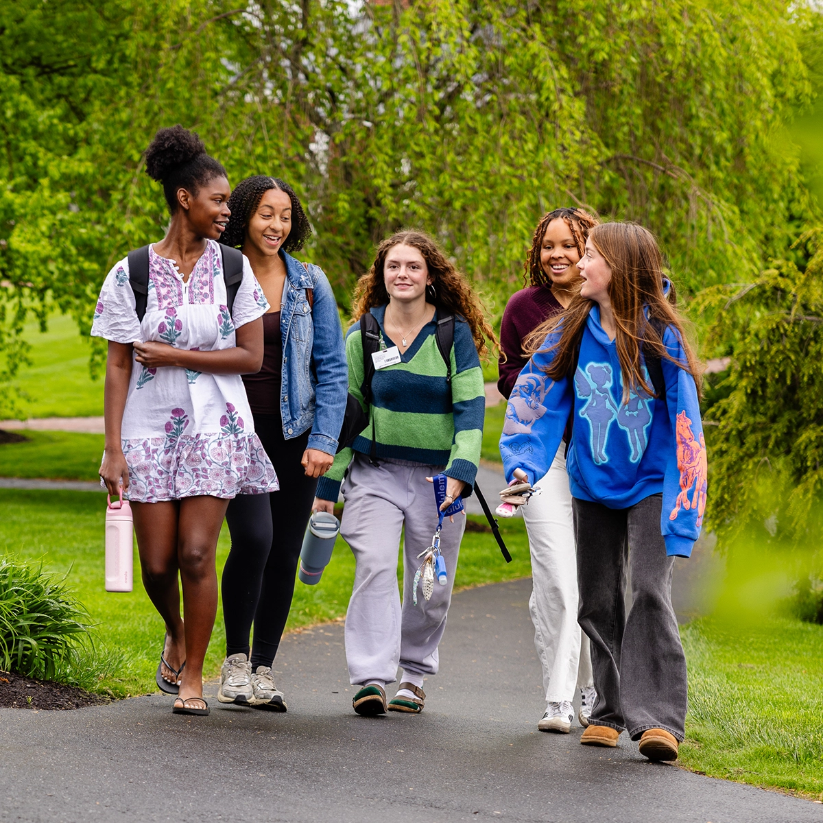 Five young women walk together on a path outdoors, smiling and talking, each carrying backpacks and water bottles, with green trees in the background.