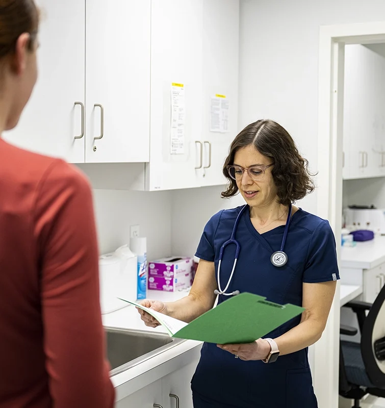A healthcare professional in navy scrubs reviews a green folder while speaking with a person in a red shirt inside a medical office with white cabinets and a sink.