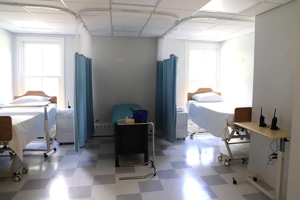 A bright hospital room with two beds separated by blue privacy curtains, a small table with a basket in the center, and medical equipment on the sides.