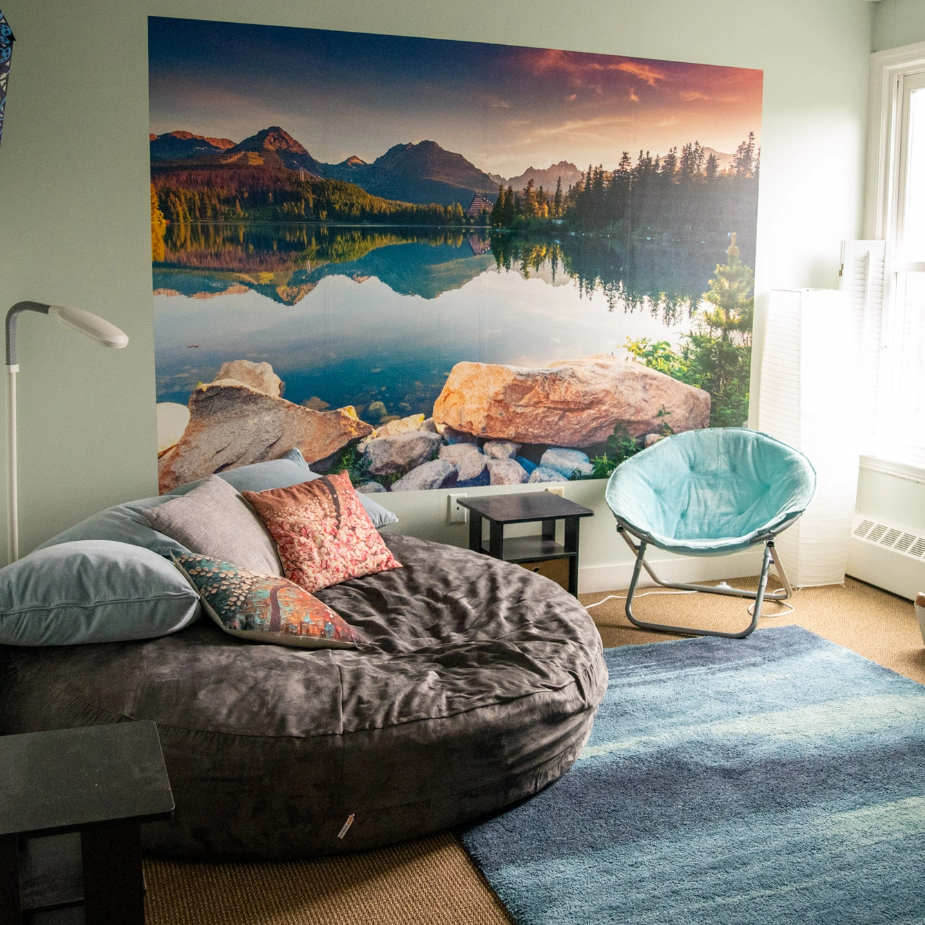 Cozy room with a large round beanbag, blue chair, small tables, floor lamp, and a scenic wall mural of a mountain lake reflecting pine trees and peaks. Blue rug covers part of the tan floor.