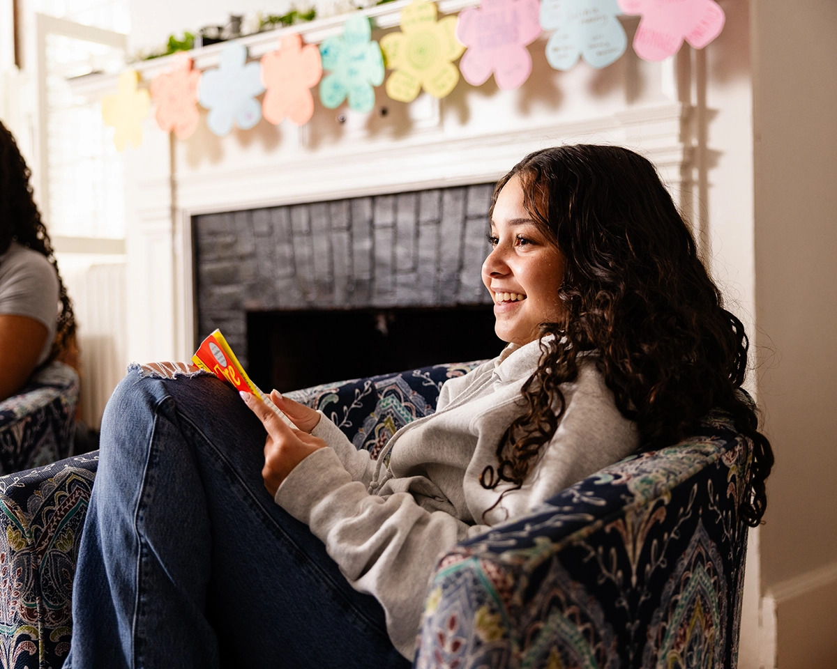 A girl with curly hair sits in an armchair, smiling and holding a colorful card. Behind her is a fireplace and a string of paper decorations hanging above the mantle.