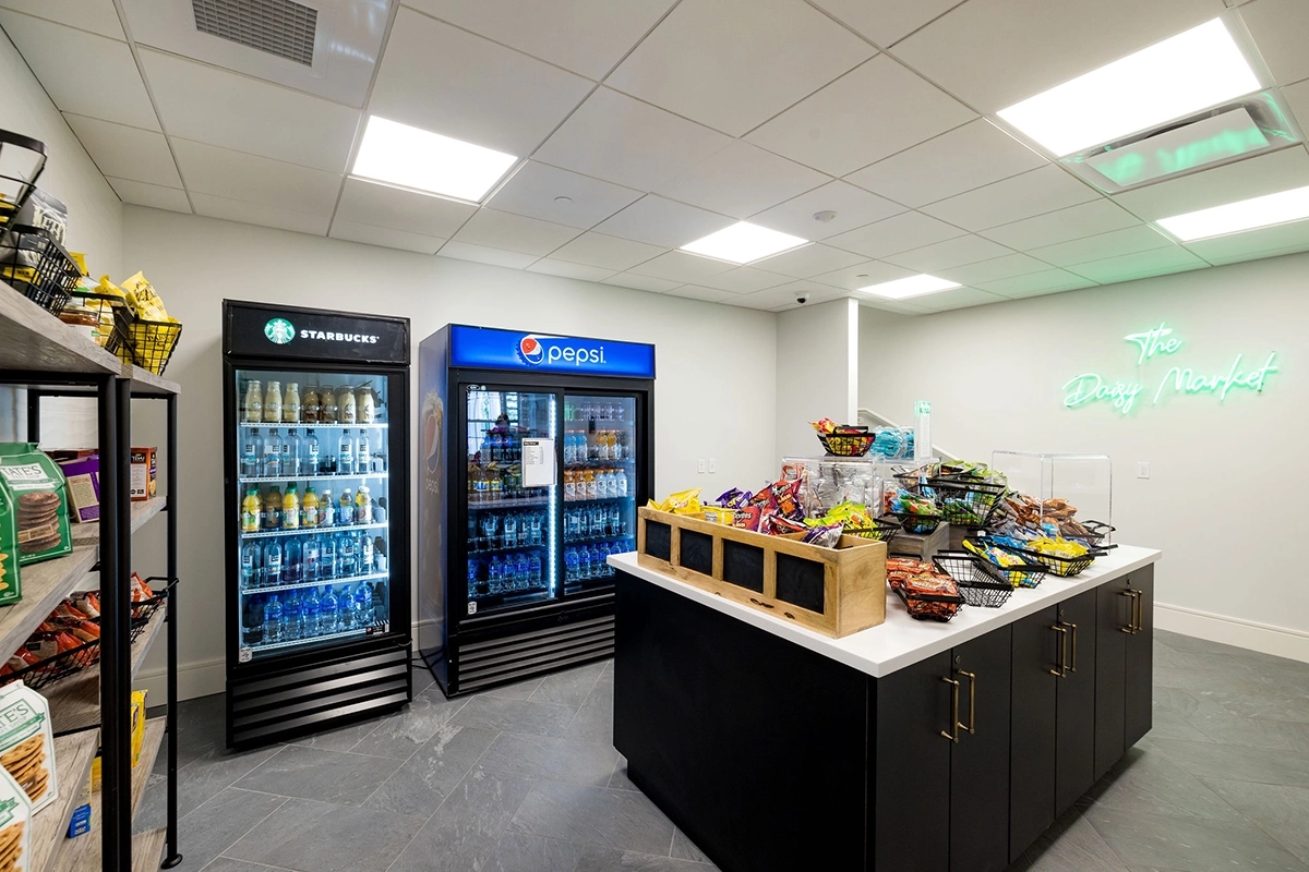 A small market features snack shelves, baskets of chips, and drink coolers labeled Starbucks and Pepsi. Bright ceiling lights and a neon sign reading “The Daily Market” illuminate the clean space.
