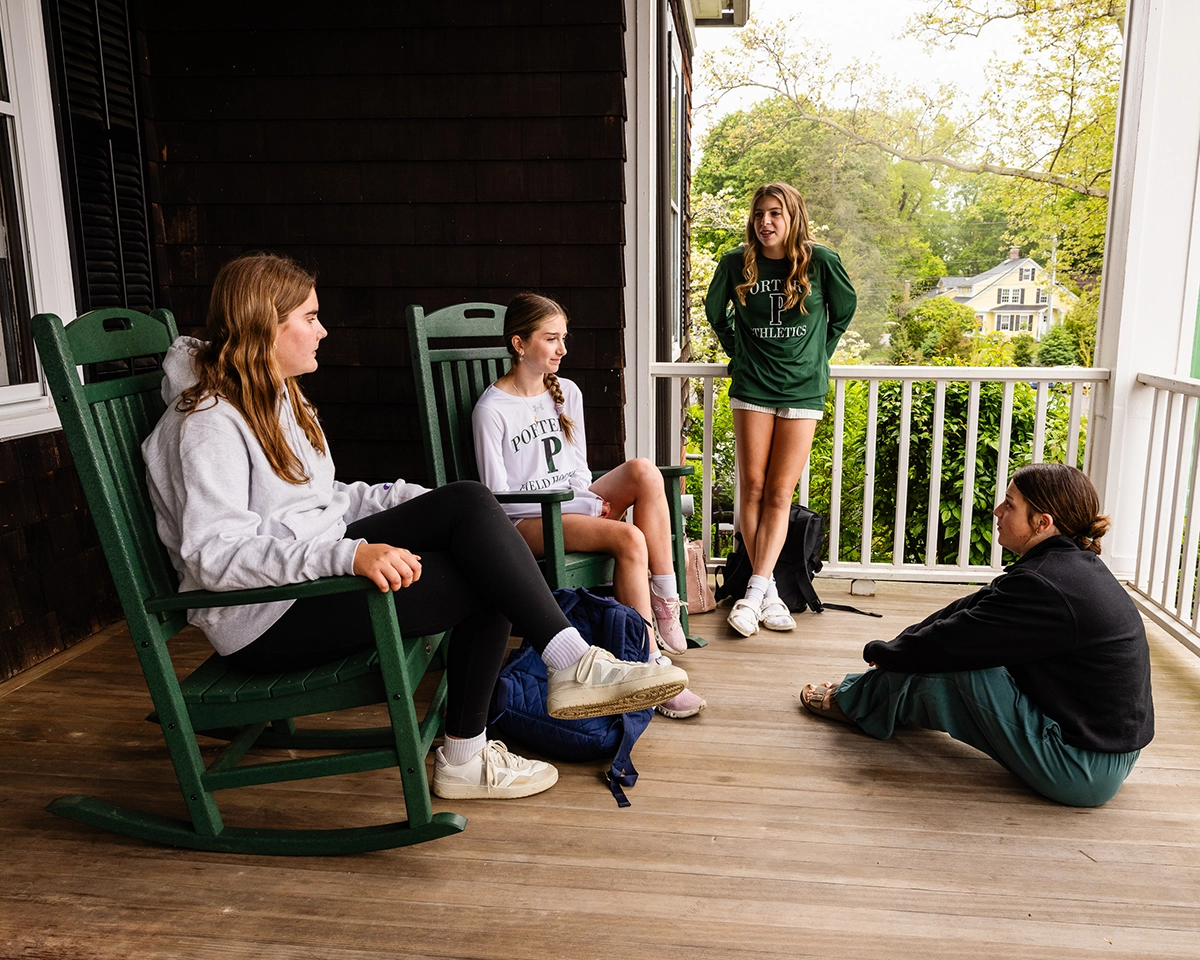 Four girls sit and stand on a wooden porch, talking. Two sit on green rocking chairs, one sits on the floor, and one stands by the railing. They wear casual clothes and the background shows trees and houses.