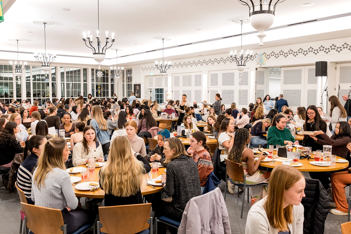 Large dining hall filled with people sitting at round tables, eating and talking. The room is brightly lit with modern chandeliers, and large windows line the walls. Most attendees appear engaged in conversations.