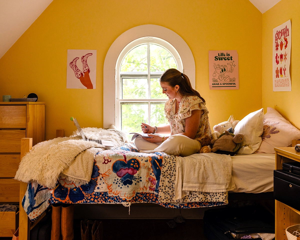 A young woman sits on her bed in a yellow-walled room, writing in a notebook with a laptop in front of her. The room has posters, stuffed animals, and sunlight coming in through an arched window.