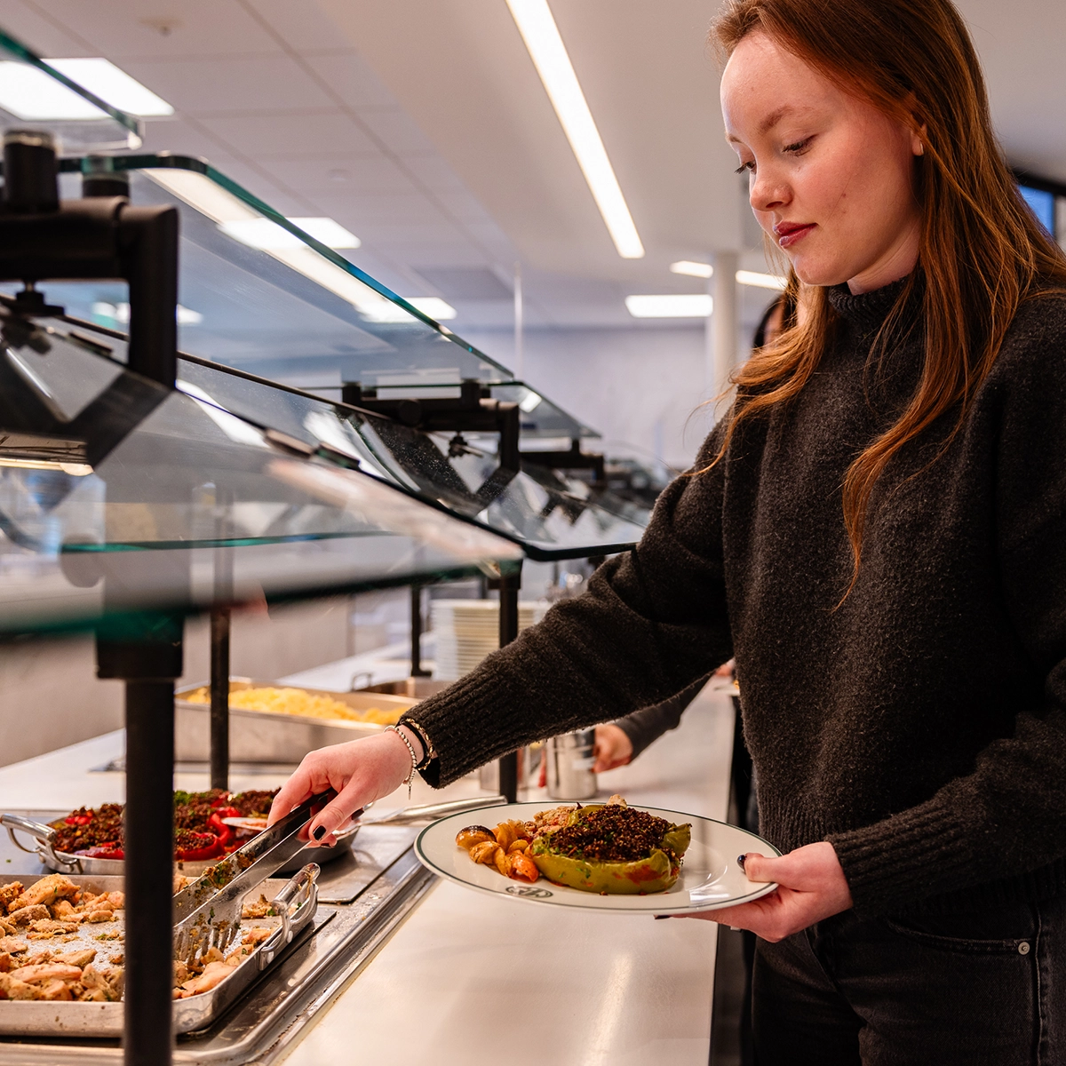 A woman serves food onto her plate from a buffet in a cafeteria. She wears a dark sweater and stands next to trays filled with various dishes under a glass sneeze guard.