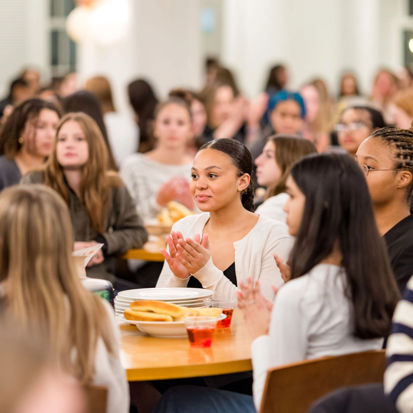 A group of people sit at round tables, some clapping and some watching attentively. The tables have plates, drinks, and food. The background shows more people, all indoors in a well-lit room.