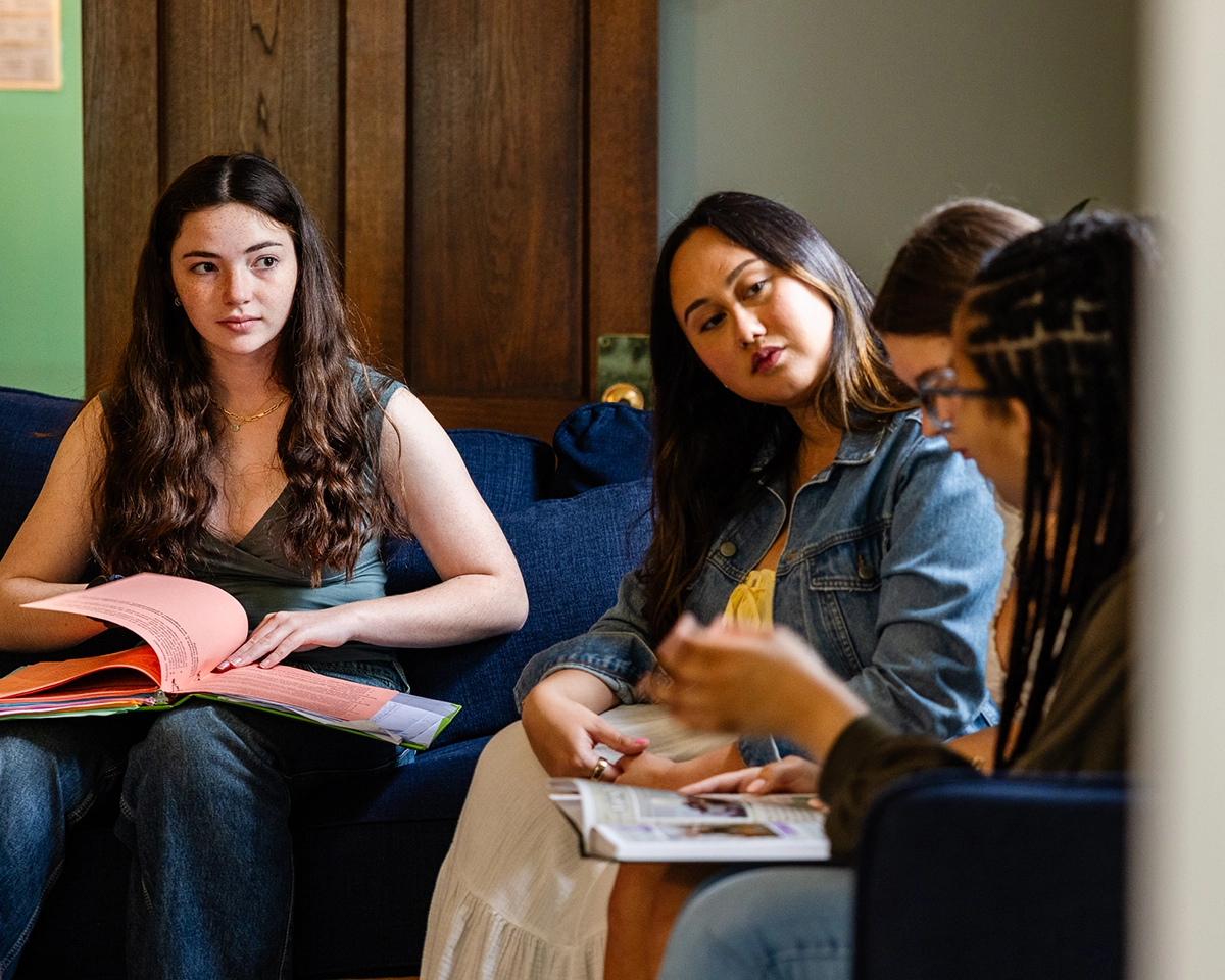 Four women sit on a blue couch having a discussion. One holds a pink folder, another writes in a notebook, and two others listen attentively. The setting appears informal and comfortable.