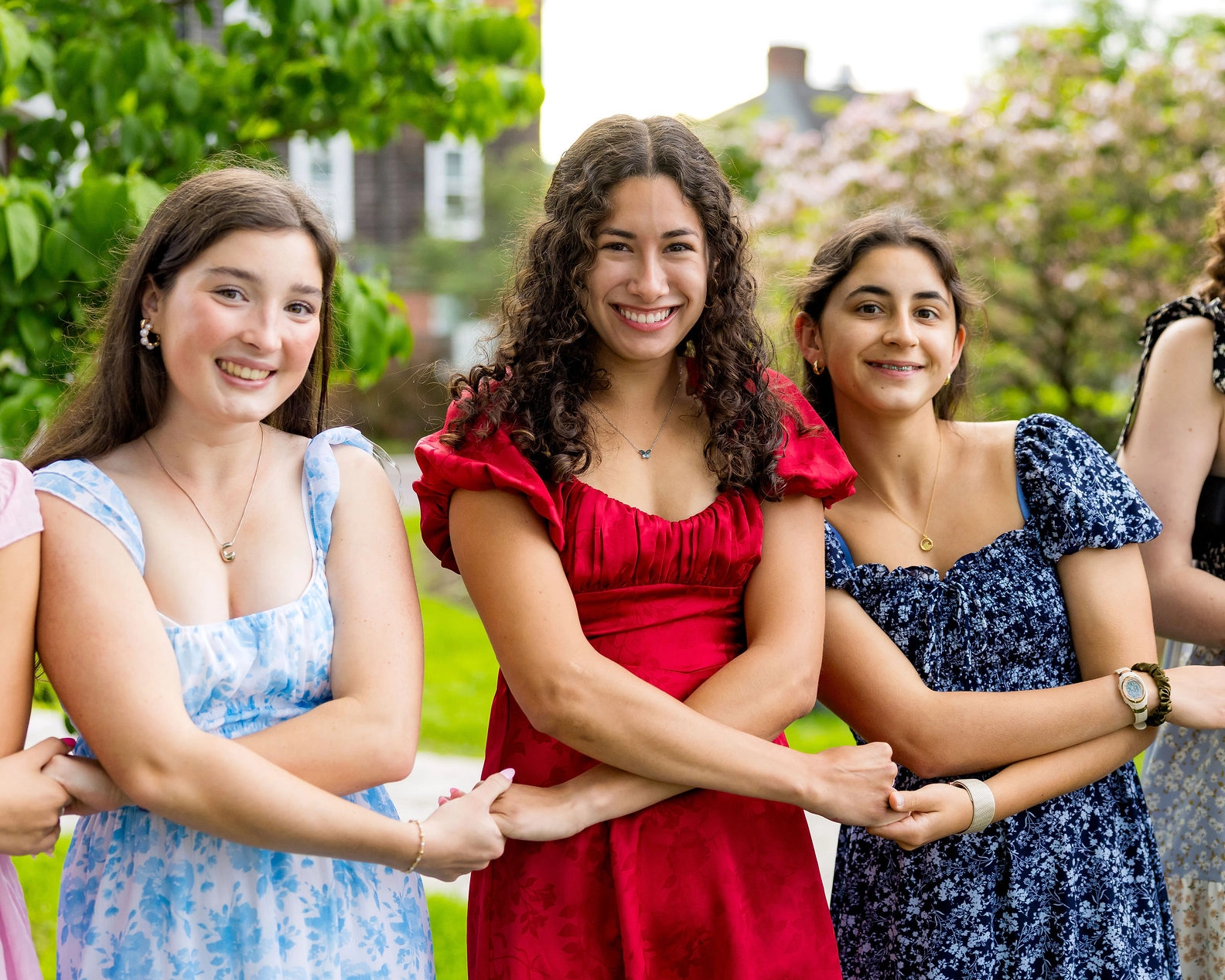 Three young women in floral dresses and a red dress stand outdoors, smiling and linking arms. Green trees and blurred buildings are visible in the background.
