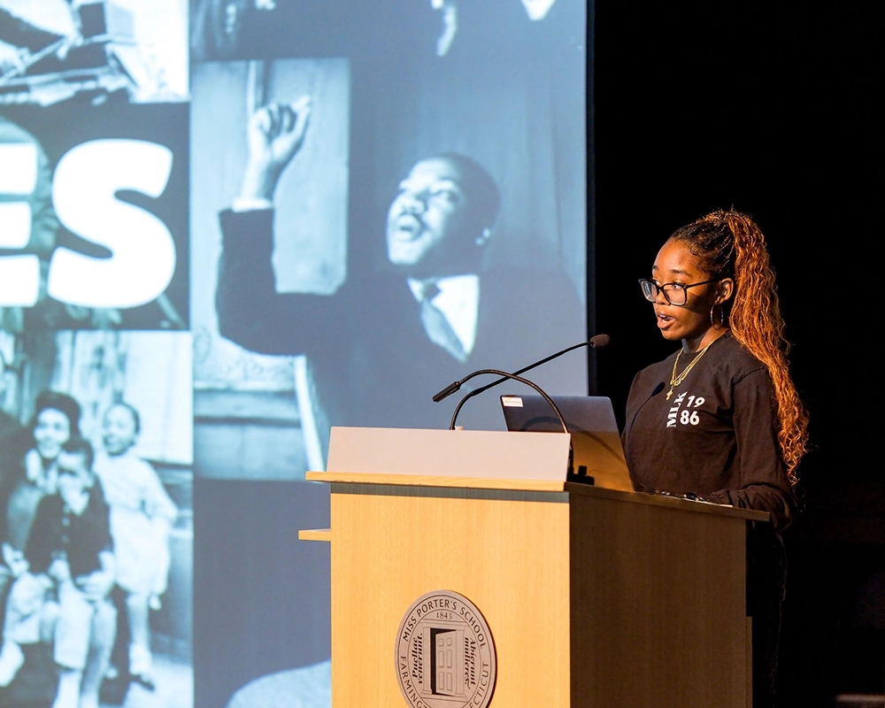 A woman speaks at a podium with a laptop, in front of a large projected image featuring a historic black-and-white photo and the word “YES.”.