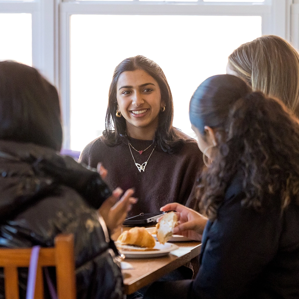 Four women sit around a table, smiling and talking. One woman faces the camera, wearing a dark sweater and a butterfly necklace. Plates with pastries are on the table, and bright windows are in the background.