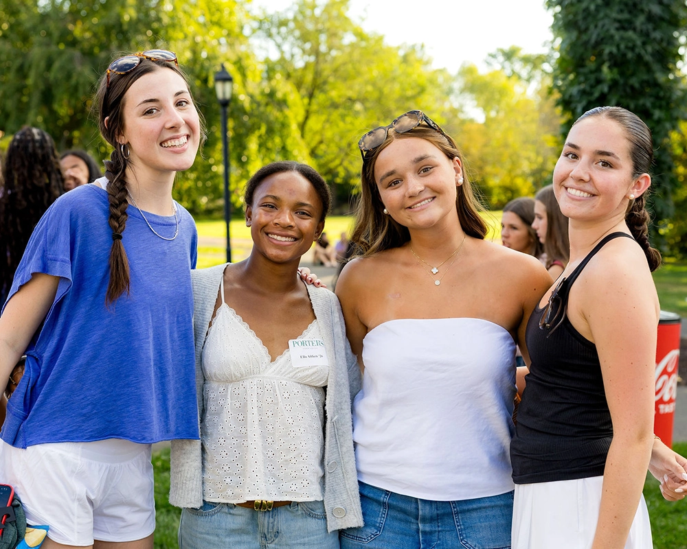Four young women standing close together outdoors, smiling at the camera on a sunny day. They are casually dressed, and trees and grass are visible in the background.