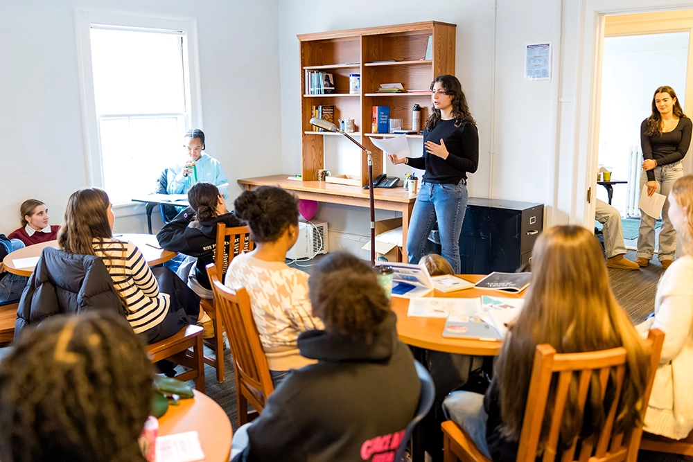 A woman stands at a podium speaking to a seated group in a classroom. People listen attentively, some taking notes. Shelves with books and papers are in the background.