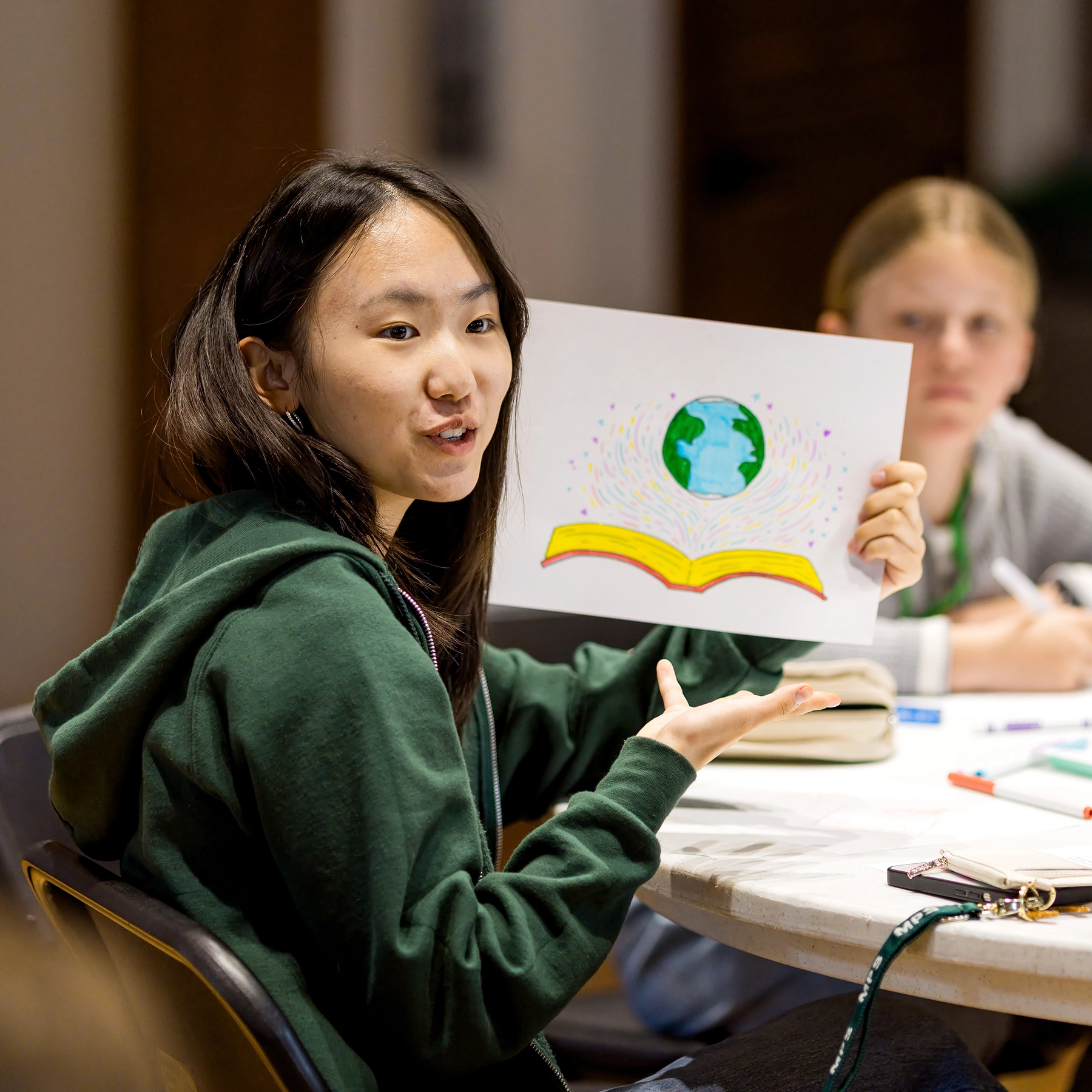 A young woman holds up a drawing of an open book with a globe above it, sitting at a table with art supplies, while another person looks on in the background.